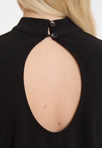 Schwarzes, ärmelloses Oberteil mit hohem Kragen und einem großen Ausschnitt auf der Rückseite, der mit zwei schwarzen Knöpfen gesichert ist. Glatte Stofftextur.