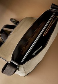 Canvas laptop bag with a beige exterior, dark brown leather accents, spacious interior, and a smooth fabric lining. Features adjustable strap and snap closure.