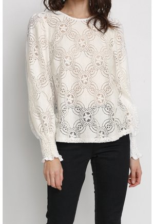 Woman wearing a white lace long-sleeve blouse with floral patterns and smocked cuffs, paired with black pants.