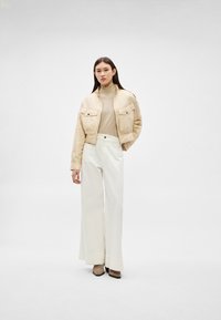 Sieviete, tērpusies bēša krāsas saīsinātā žaketē, gaiši bēšā augstā apkaklī, plaša piegriezuma baltās biksēs un pelēkbrūnās, ar punktētu purngala apaviem, kas stāv pret balto fonu.