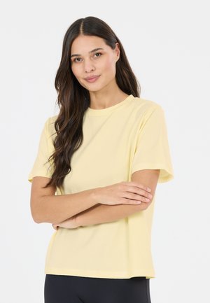 Donna con lunghi capelli scuri che indossa una maglietta gialla semplice a maniche corte, con le braccia incrociate, in piedi davanti a uno sfondo bianco uniforme.