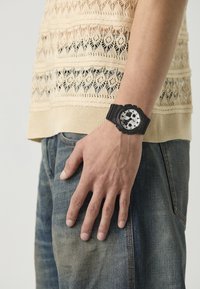 Black watch with a round face, silver accents, and a textured rubber strap. Styled on a hand against a knit beige top and denim jeans.