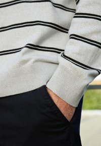 Grå tröja med svarta ränder, ribbade ärmar och mjuk textur. Bärs över mörka byxor med en hand vila i fickan.