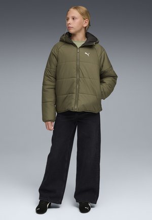 Ung person iført en olivengrøn Puma dunjakke, sorte bukser med brede ben og sorte Puma-sneakers, stående med den ene hånd i lommen.