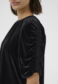 Blusa in velluto nero con maniche a sbuffo arricciate, caratterizzata da una consistenza morbida e una finitura liscia. Collo rotondo e drappeggio elegante.