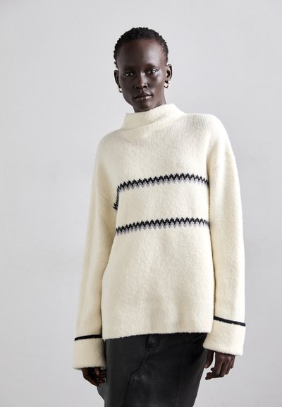 Cremefarvet sweater med høj krave, prydet med sorte zikk-zakkede striber og brede ærmer. Blødt, tekstureret stof giver varme og komfort.
