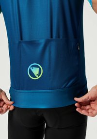Maillot de sport bleu en tissu texturé, avec une poche arrière et un logo circulaire. L'ourlet est de couleur bleue unie, conçu pour les activités de plein air.