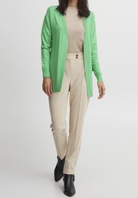 Kobieta w jasnozielonym kardiganie, beżowej bluzce, beżowych spodniach z wysokim stanem z guzikami i czarnych botkach do kostek, stojąca na białym tle.