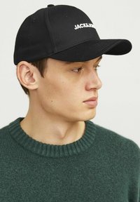 Black baseball cap with white "JACK & JONES" text, paired with a green ribbed sweater, showing a close-up profile view.