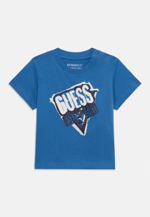 Tricou albastru pentru copii mici, cu mâneci scurte, capse cu butoni pe umăr și logo-ul "GUESS EST 1981" alb și negru pe partea din față.