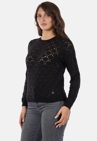 Maglione nero lavorato a maglia con motivo a diamante aperto, maniche lunghe e orlo a coste. Abbinato a jeans grigi aderenti.