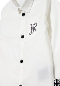 Camicia bianca con colletto a punta, bottoni neri, taschino sul lato sinistro con ricamo "JR" e motivo Union Jack sulle maniche.