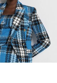 Žena v modrém, černém a bílém kostkovaném saku se zlatými knoflíky, přes odpovídající kostkovaný top, stojící proti hladké zdi.
