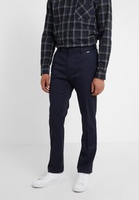 Marinblå byxor med rak passform, tillverkade av ett mjukt tyg; har bälteshällor och platt framdesign, kombinerat med vita sneakers.