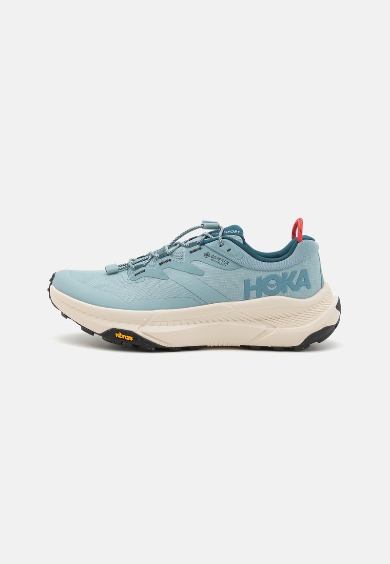 Svetlo modra tekaška čevlja z teksturirano zgornjo plastjo, oznako GORE-TEX, debelim belim srednjim podplatom in črnim Vibram podplatom. Odbijajoči detajli in zanka na zadnjem delu.