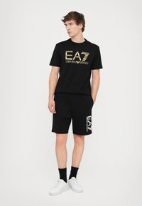 Black T-shirt with gold EA7 logo and black shorts featuring a white logo detail. Worn with white sneakers and black socks.