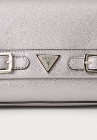 Bolso de mano plateado texturizado con un logo triangular de Guess y acentos en tono dorado. Presenta conexiones para correas y tiene una forma elegante.