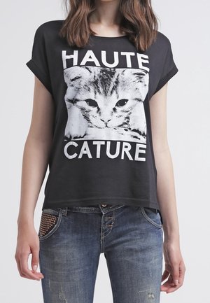 Kobieta w czarnej koszulce z dużą szarą twarzą kota i napisem "HAUTE CATURE", w połączeniu z niebieskimi dżinsami.