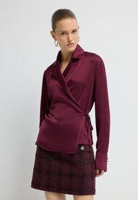 Burgundy satin omslagsbluse med lange ærmer, V-hals og drawsides, parret med en ternet teksturskørt med dekorative knapper.