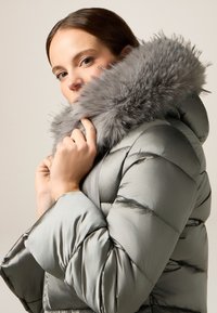 Giacca puffa grigia chiaro con colletto in pelliccia sintetica, caratterizzata da una texture trapuntata e un cappuccio caldo. Il modello tiene il colletto vicino al viso.