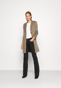 Blazer longline à carreaux marron sur un t-shirt blanc, associé à un jean flare noir et des bottines noires. Texture lisse, coupe ajustée.