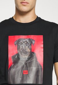 Černé tričko s grafickým potiskem rottweilera v mikině na červeném pozadí. Materiál se jeví jako lehký a měkký.