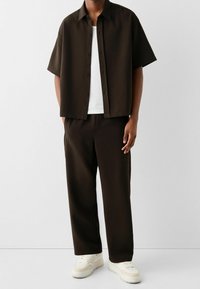 Kortärmad mörkbrun skjorta med knappstängning, kombinerad med matchande byxor med avslappnad passform och ljusfärgade sneakers. Slät textur.