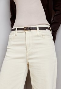 Beige høytlivte jeans med et klassisk fem-lomme design, parret med et mørkebrunt lærbelte med en metallspenne. Glatt stofftekstur.