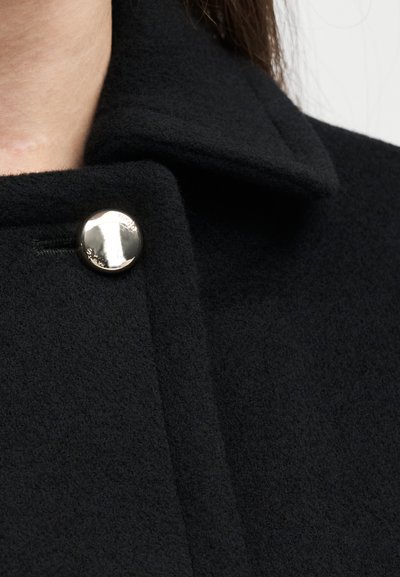 Nahaufnahme eines schwarzen Wollmantelkragens, der nahe dem Hals mit einem glänzenden, runden silbernen Knopf befestigt ist.