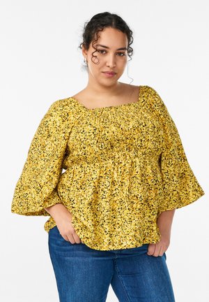 Woman with curly hair wearing a yellow floral smocked blouse with wide sleeves and blue jeans, standing with hands in pockets.