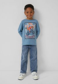 Jongentje dat glimlacht, met een blauwe longsleeve shirt met een tijger op een motorfietsafbeelding en tekst, blauwe jeans en witte sneakers.