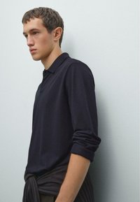 Junger Mann mit kurzen Haaren, der ein dunkles Langarm-Polohemd und einen dunklen Pullover um die Taille gebunden trägt, steht gegen eine helle Wand.