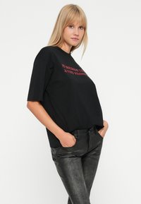Tânără îmbrăcată într-un tricou oversized negru cu imprimeu și pantaloni texturați închisi la culoare, stând cu mâinile în buzunare, pe un fundal alb.