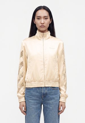 Femme aux longs cheveux noirs portant une veste zippée en satin beige avec des manches à motifs et un jean bleu clair, debout devant un fond uni.
