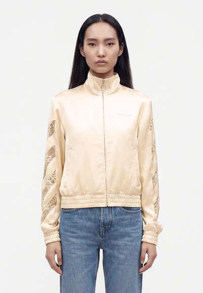 Femme aux longs cheveux noirs portant une veste zippée en satin beige avec des manches à motifs et un jean bleu clair, debout devant un fond uni.