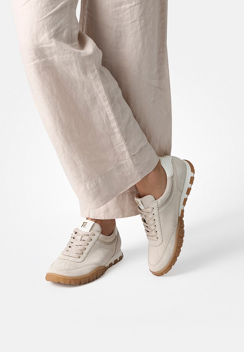 Beige sneakers with textured fabric, lace-up design, and rubber sole featuring a brown tread. Accented with a white heel and logo detail.