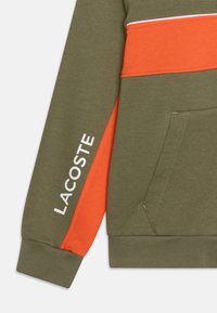 Hoodie vert olive avec un panneau orange, accents blancs, et le mot "LACOSTE" imprimé en blanc sur le côté. Texture lisse, poche kangourou.
