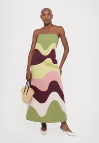 Strapless maxi dress in green, pink, and burgundy wave patterns. Smooth fabric with a flowy design. Model holds a woven bag and wears white shoes.