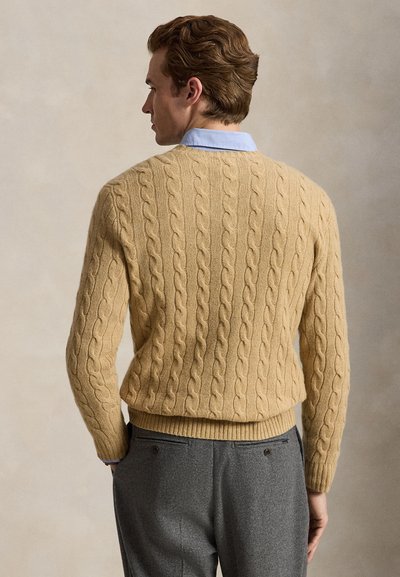 Hombre con el cabello corto y castaño de espaldas, lleva un suéter beige de punto con diseño de cables sobre una camisa de cuello azul claro y pantalones grises.