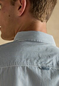 Koszula w pionowe niebieskie i białe cienkie paski, ze standardowym kołnierzykiem i małą metką logo z tyłu kołnierza, o gładkiej teksturze materiału.