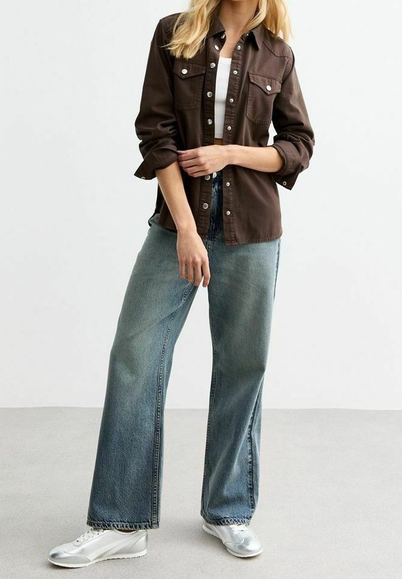 Femme portant une chemise marron foncé boutonnée sur un haut blanc, un jean large bleu clair et des baskets argentées, debout devant un fond uni.