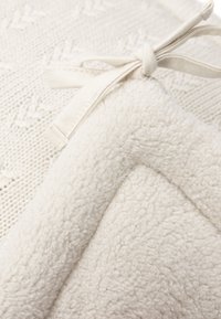 Couverture de couleur crème avec une texture en tricot d'un côté et en polaire douce et pelucheuse de l'autre. Comprend un lien en ruban pour la fermeture.