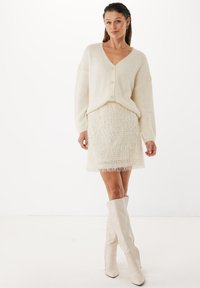 Krämfärgad, fluffig cardigan med V-ringning och knappar, i kombination med en texturerad, fransad minikjol. Modellen bär höga, krämfärgade, spetsiga stövlar.