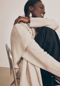 Maglione beige con design drappeggiato e dettagli a coste, abbinato a pantaloni neri. Presenta grandi orecchini di perle e una sedia in metallo elegante.