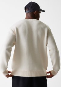 Pulover tricotat de culoare crem, cu un finisaj texturat, tiv și manșete cu rib. Pur si simplu, este purtat invers, evidențiind un design simplu și casual.