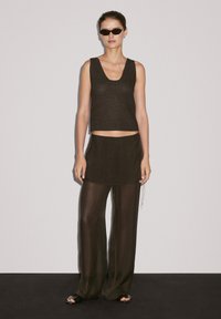 Crop top noir en tricot sans manches avec une encolure dégagée, associé à une jupe semi-transparente marron foncé et un pantalon large assorti. Sandales noires.