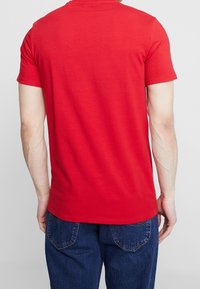 Rød bomulds T-shirt med korte ærmer og rund halsudskæring, vist fra ryggen. Bæres med blå denimjeans, der har baglommer.