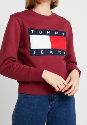 Kobieta ubrana w bordową bluzę z dużą naszywką z logo Tommy Jeans, połączoną z niebieskimi dżinsami, ręce splecione na wysokości talii.