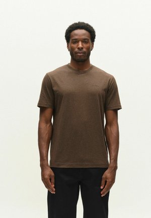 Mand med kort krøllet hår iført en ensfarvet brun kortærmet t-shirt og sorte bukser, der står mod en lys baggrund.