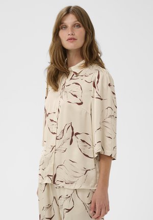 Blouse beige clair à motif de feuilles marron, manches courtes, boutonnée sur le devant, avec col. Tissu lisse avec une coupe décontractée.